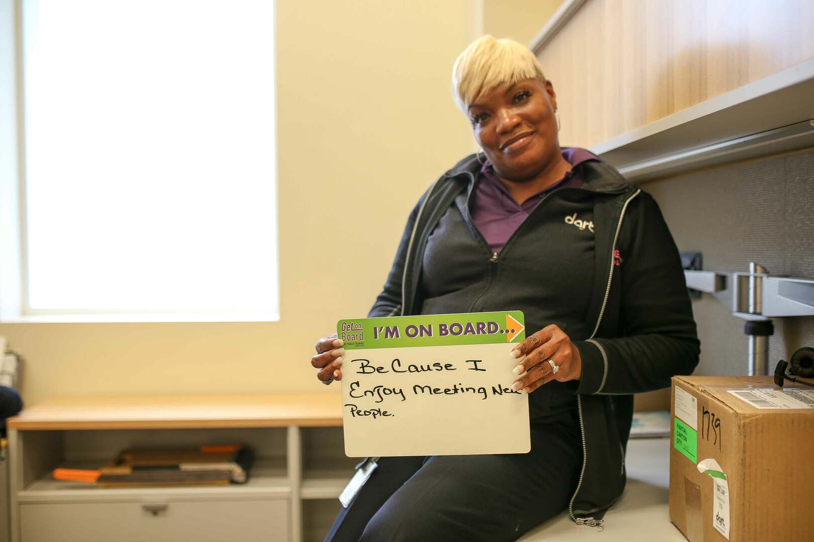 Woman rests against desk hold sign reading "I'm on board because I enjoy meeting new people."