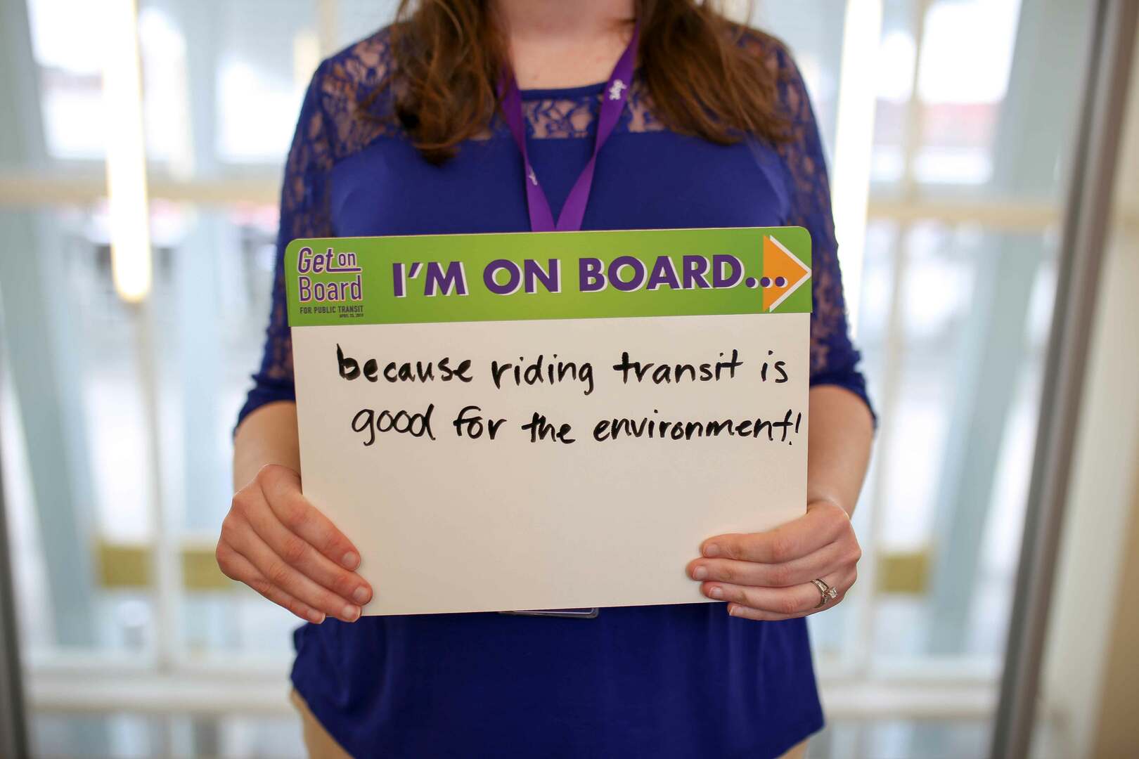 Woman in blue shirt poses with sign reading "I'm on board because riding transit is good for the environment."